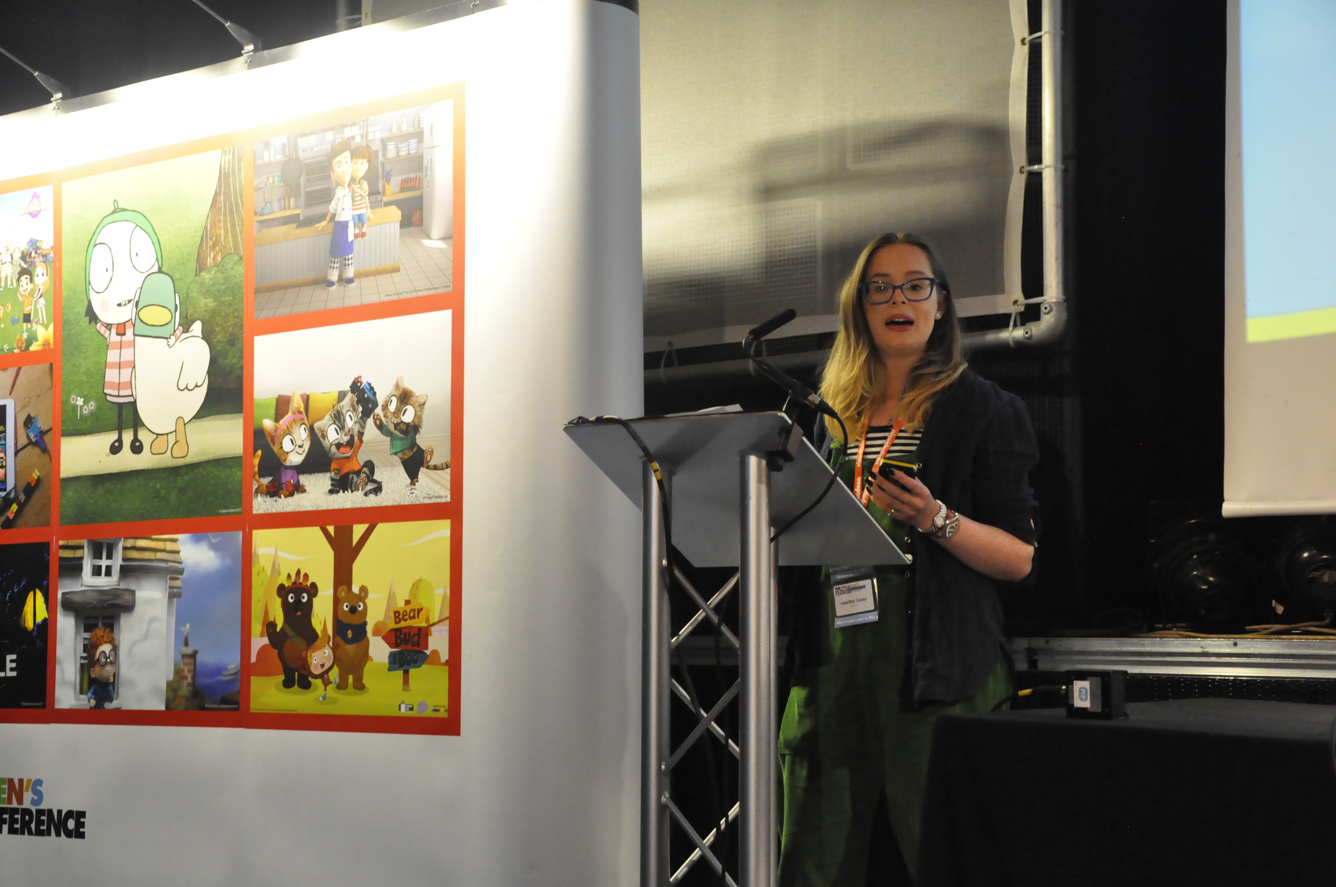 A woman stood at a lectern wearing green dungarees pictured mid-sentence. Next to her is a large board covered in pictures and there's a large screen on the right of the picture with a powerpoint presentation 