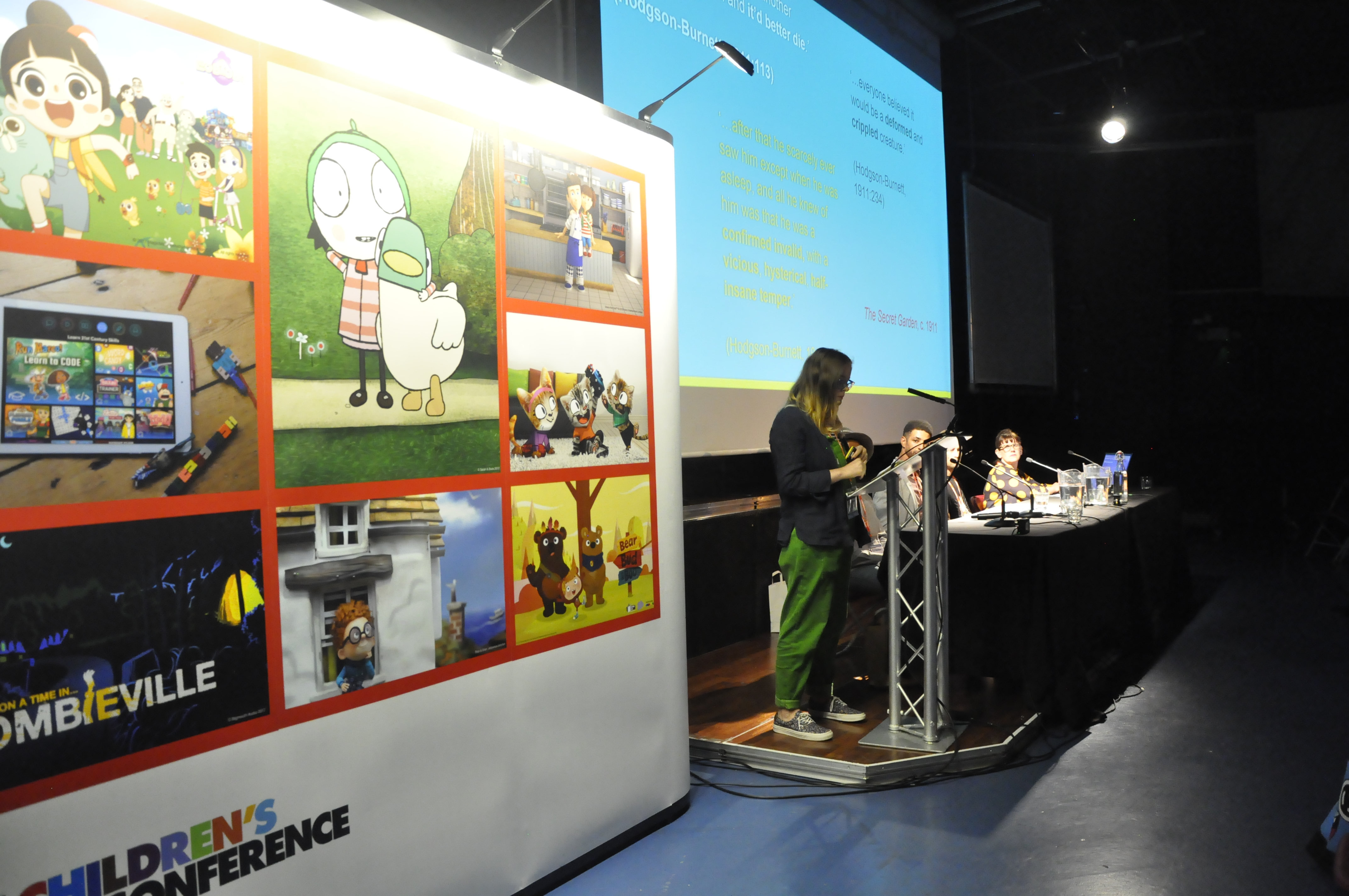 A woman stood at a lectern wearing green dungarees. You cannot see the front of her face. Next to her is a large board covered in pictures and there's a large screen on the right of the picture with a powerpoint presentation.  
